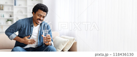 African American man is seated on a light-colored couch, holding a glass of water while showing signs of discomfort. He appears to be in a living room with bright, soft lighting. African American man is seated on a light-colored couch, holding a glass of water while showing signs of discomfort. He appears to be in a living room with bright, soft lighting. 118436130