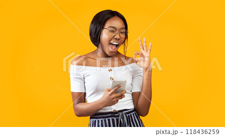 Great App. Positive Afro Girl Holding Phone Gesturing Okay And Winking At Camera Standing Over Orange Studio Background. Panorama Great App. Positive Afro Girl Holding Phone Gesturing Okay And Winking At Camera Standing Over Orange Studio Background. Panorama 118436259