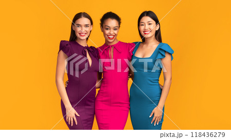 It Girls. Three Diverse Young Ladies In Cocktail Dresses Smiling At Camera And Hugging Standing On Yellow Background. Studio Shot 118436279