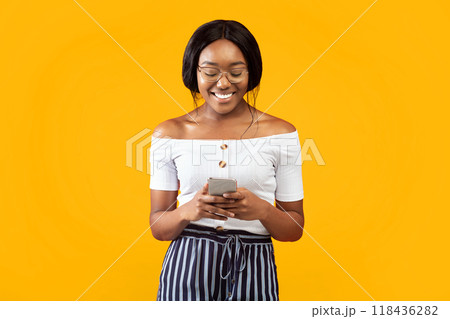 Smiling Black Lady Using Smartphone Chatting With Friends In Social Media Standing Over Orange Background. Studio Shot Smiling Black Lady Using Smartphone Chatting With Friends In Social Media Standing Over Orange Background. Studio Shot 118436282