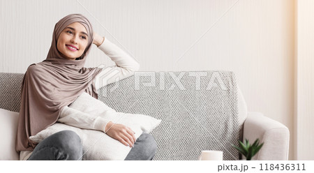 Daydreaming. Relaxed arabic girl in hijab sitting on sofa in stylish interior, thinking about something, enjoying staying at home 118436311