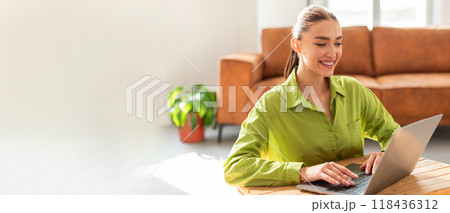 A woman with long hair smiles while typing on her laptop at a wooden table. The well-lit living room features a cozy couch and a green plant, creating a welcoming atmosphere. 118436312