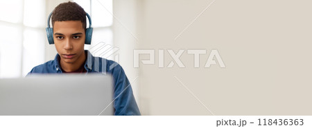 A young African American man is deeply engaged in his computer work, wearing headphones. The bright room features large windows that provide natural light, enhancing his concentration. 118436363