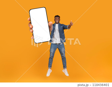 A joyful young African American man is leaping in the air, holding a smartphone towards the camera while giving a thumbs up. The vibrant orange background highlights his excitement and energy. 118436401