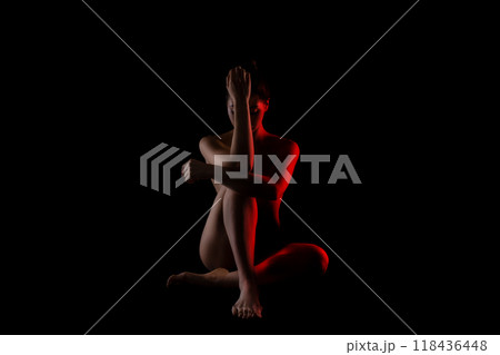 The image shows a woman sitting in a dark studio with red light illuminating her body. She is facing the camera with her hands clasped together near her chest, and her head is slightly tilted down The image shows a woman sitting in a dark studio with red light illuminating her body. She is facing the camera with her hands clasped together near her chest, and her head is slightly tilted down 118436448
