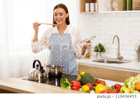 A happy woman in an apron tastes food while cooking in her kitchen. She is surrounded by fresh vegetables and cooking supplies. A happy woman in an apron tastes food while cooking in her kitchen. She is surrounded by fresh vegetables and cooking supplies. 118436453