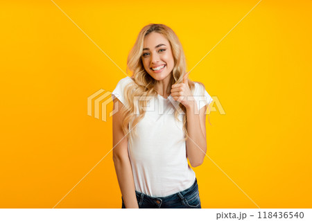 A young woman with long, blonde hair smiles and gives a thumbs up while standing in front of a bright yellow background. She is wearing a white t-shirt and blue jeans. 118436540