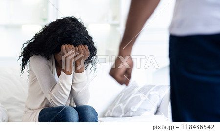 A woman sits on a couch, crying with her head in her hands, while a man stands behind her. 118436542