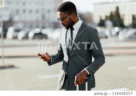 African American man in a suit is using his phone while standing with luggage near an airport entrance. The setting is modern and professional, highlighting business travel. 118436557