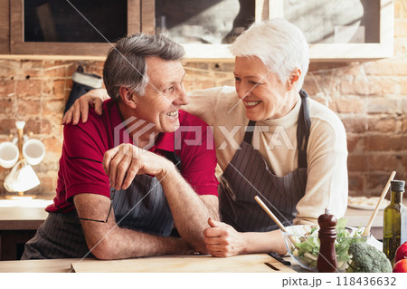 A senior couple smiles at the camera while standing in their modern kitchen. The man is wearing a red shirt and an apron, and the woman is wearing a beige sweater and an apron A senior couple smiles at the camera while standing in their modern kitchen. The man is wearing a red shirt and an apron, and the woman is wearing a beige sweater and an apron 118436632