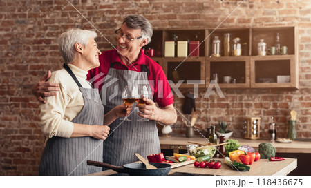An older couple, both wearing aprons, are standing in their kitchen, smiling and toasting with glasses of wine. The kitchen has brick walls and wooden cabinets. 118436675