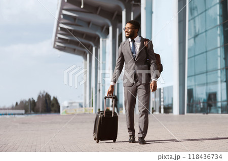 African American businessman is walking with his luggage outside a modern airport. The mood is dynamic and professional, highlighting travel and business with a sleek airport backdrop. 118436734