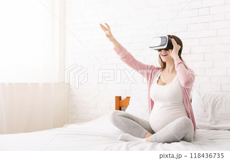 Expecting woman is sitting on a bed, wearing a virtual reality headset. She appears engaged in the virtual experience, with a focused expression on her face, copy space Expecting woman is sitting on a bed, wearing a virtual reality headset. She appears engaged in the virtual experience, with a focused expression on her face, copy space 118436735