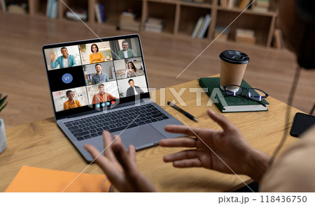 A person is sitting at a desk in front of a laptop computer, participating in a video conference call. The laptop screen displays nine people in separate video squares. 118436750