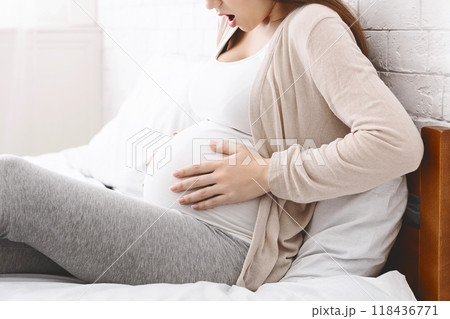 A pregnant woman sits in a bed, wearing comfortable grey pants and a light sweater, tenderly holding her belly during the afternoon. She appears to be in a relaxed home setting, cropped A pregnant woman sits in a bed, wearing comfortable grey pants and a light sweater, tenderly holding her belly during the afternoon. She appears to be in a relaxed home setting, cropped 118436771