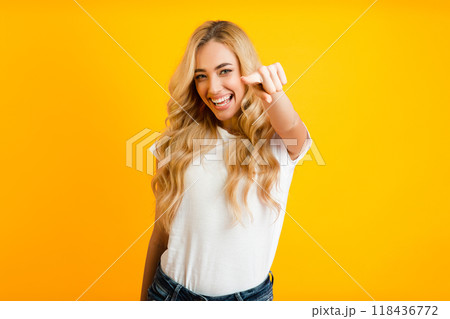 A young woman with long, blonde hair is smiling and pointing directly at the camera. She is wearing a white t-shirt and jeans and is standing in front of a solid yellow background. A young woman with long, blonde hair is smiling and pointing directly at the camera. She is wearing a white t-shirt and jeans and is standing in front of a solid yellow background. 118436772
