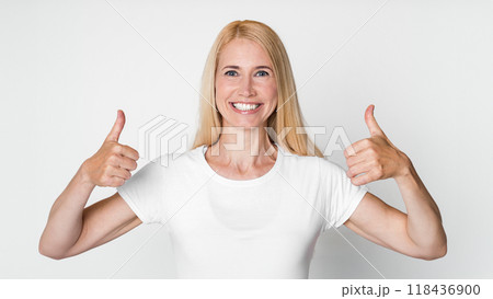 A photo of a smiling blonde woman wearing a white t-shirt against a plain white background. She is giving a thumbs up with both hands. Her smile is genuine and inviting 118436900