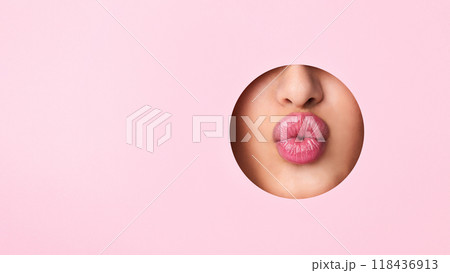 A close-up view of a woman lips protruding out of a hole against a vibrant pink background. The lips appear to be puckered or in a kissing gesture. A close-up view of a woman lips protruding out of a hole against a vibrant pink background. The lips appear to be puckered or in a kissing gesture. 118436913