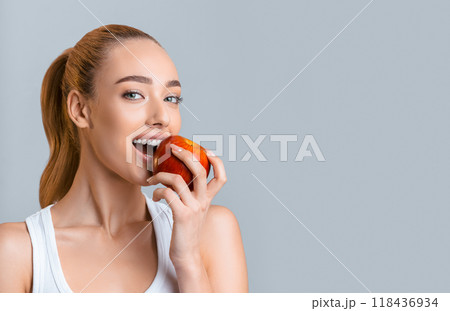 A young woman with long blonde hair tied back in a ponytail smiles as she bites into a red apple. She is wearing a white tank top and is standing in front of a gray background, copy space 118436934