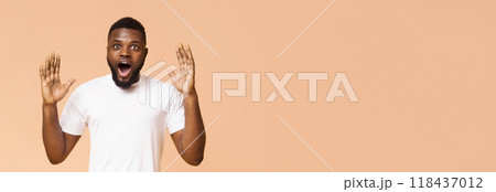 African American young man with short hair and a white t-shirt looks surprised with his mouth open and hands raised in front of a beige background, panorama with copy space 118437012