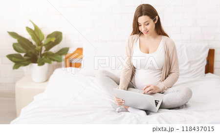 A pregnant woman sits on a bed, focusing on her laptop screen. She appears comfortable and engaged, possibly working or researching. The soft lighting in the room creates a warm and cozy atmosphere. 118437015
