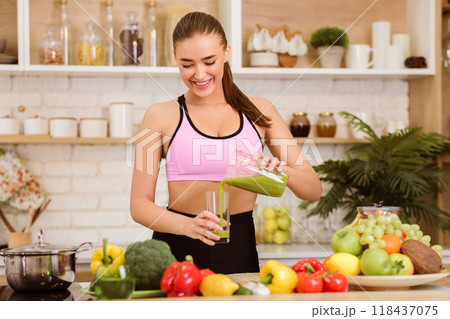A woman in a pink sports bra pours a green smoothie from a pitcher into a glass in a kitchen. 118437075
