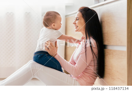 A mother with long dark hair and her baby sit on the kitchen floor, smiling and engaging in playful interaction. The bright, modern kitchen setting with ample natural light A mother with long dark hair and her baby sit on the kitchen floor, smiling and engaging in playful interaction. The bright, modern kitchen setting with ample natural light 118437134