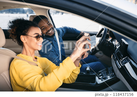 African American woman in sunglasses sits in the passenger seat of a car and takes a selfie with her boyfriend who is driving, both of them smiling at the camera. African American woman in sunglasses sits in the passenger seat of a car and takes a selfie with her boyfriend who is driving, both of them smiling at the camera. 118437156