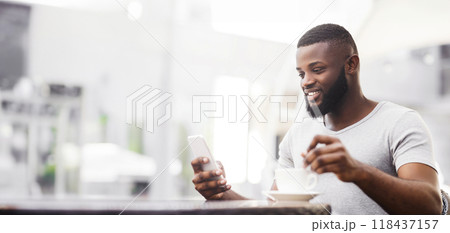 A man sits at a cafe table, smiling as he uses his phone. He is holding a cup of coffee in his other hand. 118437157