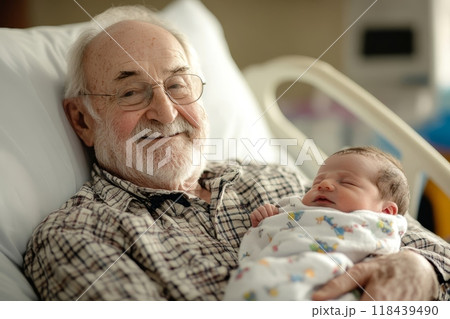 A smiling elderly man with white hair and beard holds a sleeping newborn baby in a hospital bed, capturing a heartwarming moment between generations 118439490