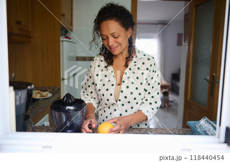 Woman preparing fresh citrus juice in cozy kitchen with sunny window, exuding warmth and homely atmosphere 118440454