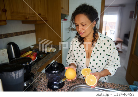 Woman preparing fresh orange juice in a cozy kitchen as she holds and squeezes ripe citrus fruits into a juicer Woman preparing fresh orange juice in a cozy kitchen as she holds and squeezes ripe citrus fruits into a juicer 118440455