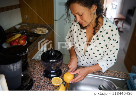 Woman preparing fresh lemon juice in a cozy kitchen with fruits on the counter and a juicer, showcasing a healthy and natural lifestyle. 118440456
