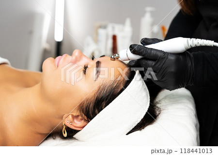 Close-up portrait of a beautiful woman with closed eyes and apparatus for skin rejuvenation. Beauty salon procedures. RF lifting. Microcurrents. 118441013