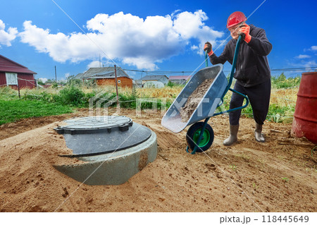 Installing wastewater plumbing for country house, worker uses wheelbarrow to transport sand. Installing wastewater plumbing for country house, worker uses wheelbarrow to transport sand. 118445649