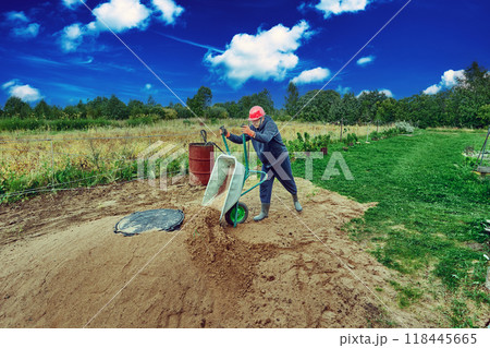Worker pours sand from wheelbarrow onto concrete ring of septic tank. 118445665