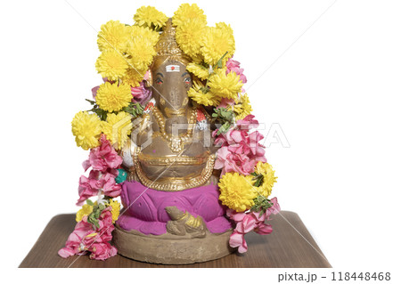 Hindu god Ganesha idol made of clay on wooden table on white background decorated with flower garlands at home in celebration of Ganesha Chaturthi festival. Closeup Hindu god Ganesha idol made of clay on wooden table on white background decorated with flower garlands at home in celebration of Ganesha Chaturthi festival. Closeup 118448468