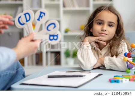 Cute girl sitting at the table while having a speech therapy tutorial and smiling nicely Cute girl sitting at the table while having a speech therapy tutorial and smiling nicely 118450954