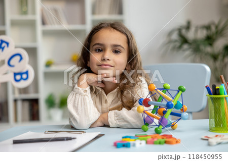 Cute girl sitting at the table while having a speech therapy tutorial and smiling nicely 118450955