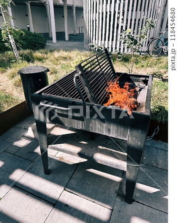 Firewood burns in an open grill in the yard against the backdrop of a modern structure, a trash can, and green grass. 118454680