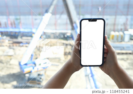 Close up of hands holding smartphone with white empty screen on blurred construction site background Close up of hands holding smartphone with white empty screen on blurred construction site background 118455241