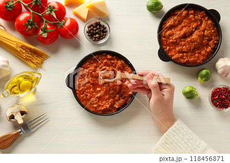 Pans with Bolognese sauce, wooden spoon in hand and ingredients on white wooden background, top view 118456871