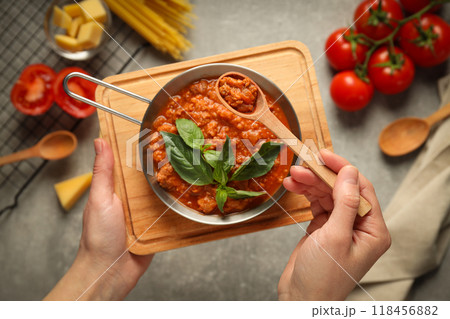Bolognese sauce in pan and spoon in hands, ingredients on gray background, top view Bolognese sauce in pan and spoon in hands, ingredients on gray background, top view 118456882