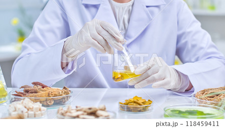 Scientist carefully holds and observes flask filled with yellow liquid, surrounded on the table by herbal ingredients contained in transparent petri dishes. Sample image for experiments in lap setting 118459411