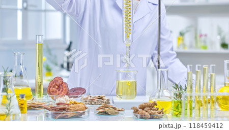 A scientist wearing a lab coat, gloves and a protective mask is holding a flask filled with a yellow liquid, examining it in a lab surrounded by herbs, flasks and other lab equipment. A scientist wearing a lab coat, gloves and a protective mask is holding a flask filled with a yellow liquid, examining it in a lab surrounded by herbs, flasks and other lab equipment. 118459412