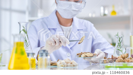 Laboratory scene featuring a scientist wearing a white lab coat and gloves using tweezers to pick up a piece of dried herb, surrounded by other dried herbs, roots, and various other lab equipment. 118460836