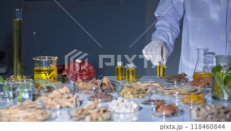 Modern lab setting with gray background features a scientist carefully picks up a vial containing a yellow liquid. The lab table is filled with various herbs, roots, and lab equipment. 118460844