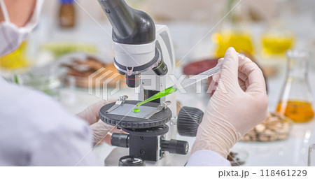 Rear view of a scientist dropping green extract onto a microscope for observation. Laboratory setting with herbs and instruments on the table, suitable for experimental photography. 118461229