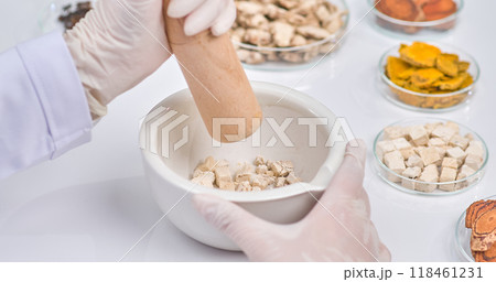 A scientist hand with a wooden pestle grinds herbal pieces in a white mortar. Surrounding ingredients showcase a vibrant array of natural herbs in petri dish. 118461231