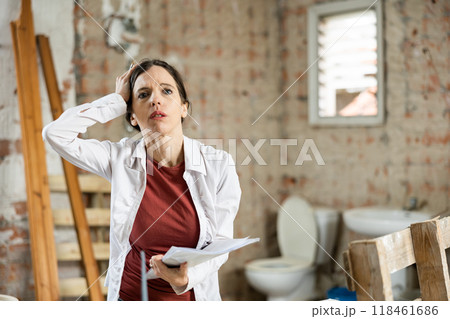 Confused female designer standing with papers in apartment during renovation Confused female designer standing with papers in apartment during renovation 118461686